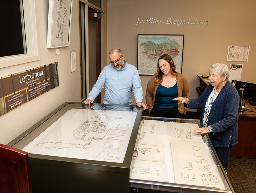 Three people stand at display cases near the entrance of the Jon Bilbao Basque Library, viewing rubbings of Basque arborglyphs laid out in glass‑covered drawers.